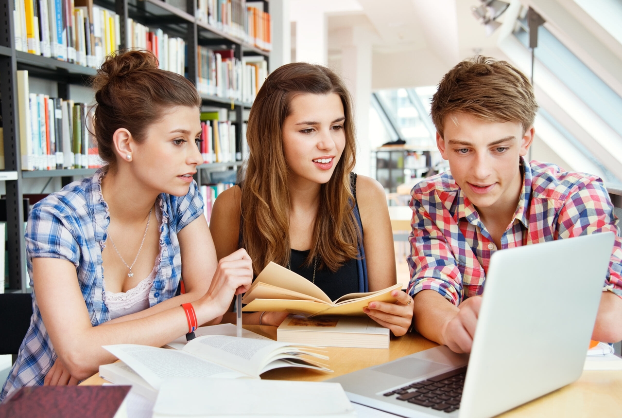 A group of students are sitting down and studying together in a library setting. They’ve got books open and are looking at a laptop.