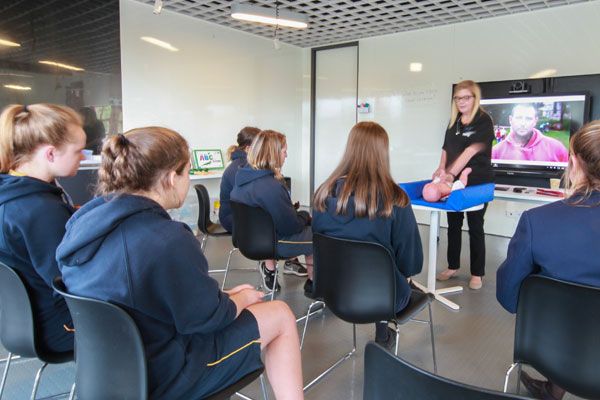 A group of students seated in chairs, attentively watching a teacher conduct a presentation in a classroom setting.