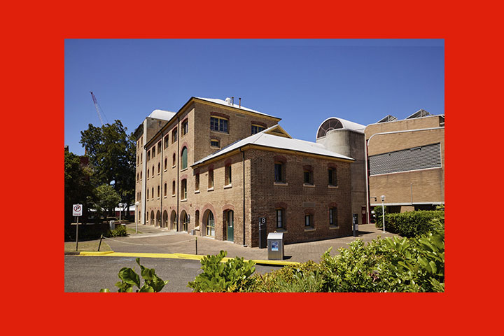TAFE NSW Hamilton campus featuring multiple heritage‑style brick buildings with arched windows and metal roofing, arranged around a paved courtyard with greenery in the foreground, set against a clear blue sky.