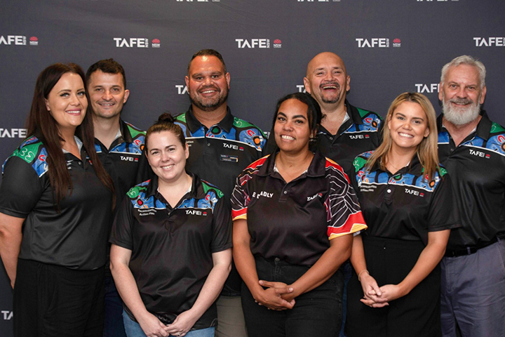 A group of eight Aboriginal TAFE NSW workers are smiling for the camera, wearing TAFE NSW shirts with a range of colourful Indigenous designs.