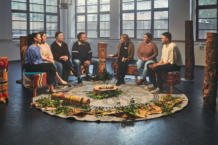 An Indigenous mob sitting in a yarning semi circle, engaging in conversation. There is a round rug that they are sitting on around, leaves, plants and other culturally significant pieces. 