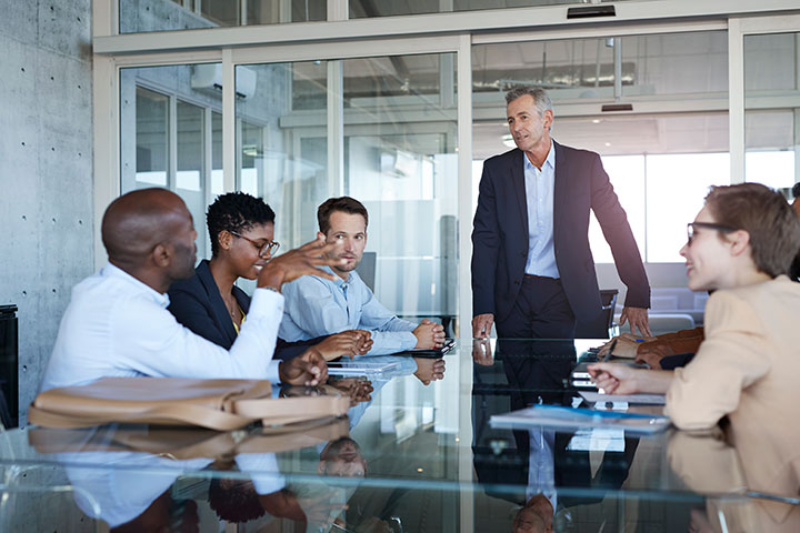 The image shows a group of professional corporate workers sitting around an office table. They are engaged in, and listening intently to someone speaking. The office is light and modern.