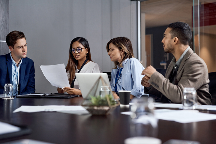 Two women and two men in formal office attire engaged in a group discussion at a boardroom table, reviewing a document together.