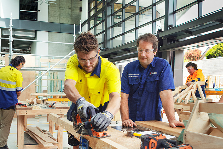 A male student, guided by his teacher, works on a carpentry project in a workshop. Two other students work on their projects in the background.