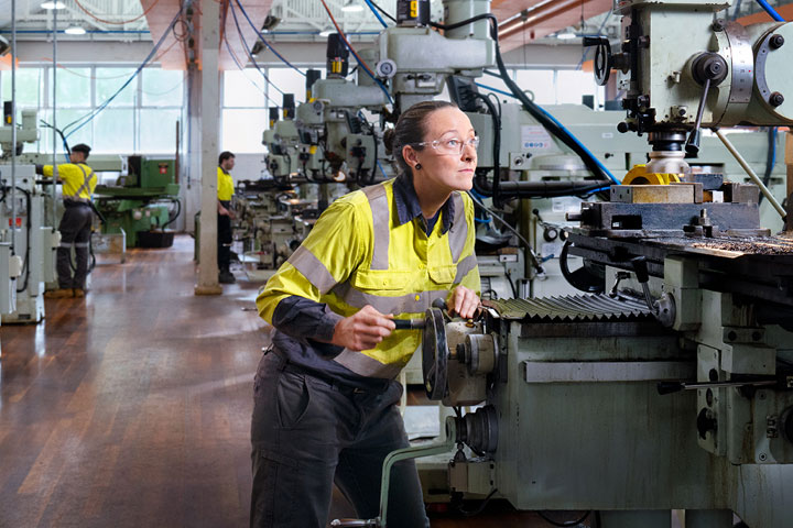 A busy workshop filled with milling machines and metal lathes. In the foreground, a woman in full PPE is closely adjusting the settings on a vertical milling machine.