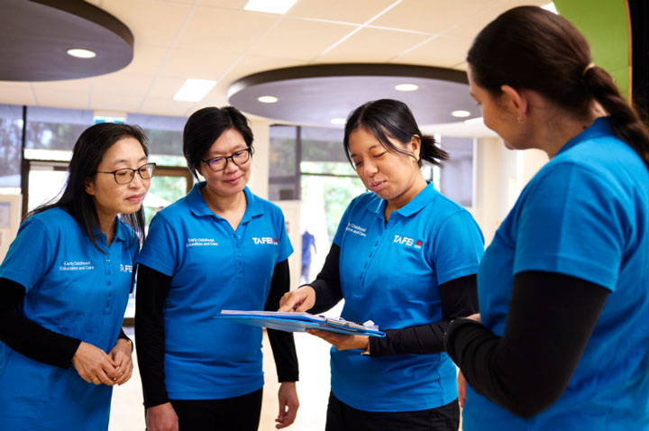 A group of four female childcare workers stand in a workspace. They all wear the TAFE NSW blue t-shirt uniform. One of the workers holds a folder while pointing out some notes to the others. They are relaxed and smiling.