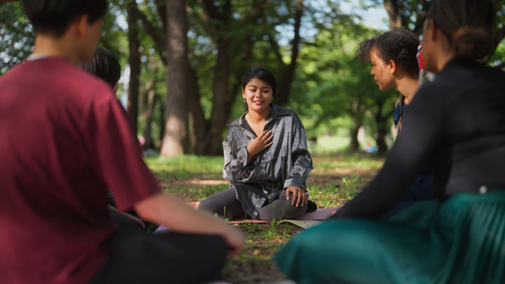 A group of people sitting on the ground in a park, all with their legs crossed. All members of the circle are looking towards one women, whom is running the circles discussion.