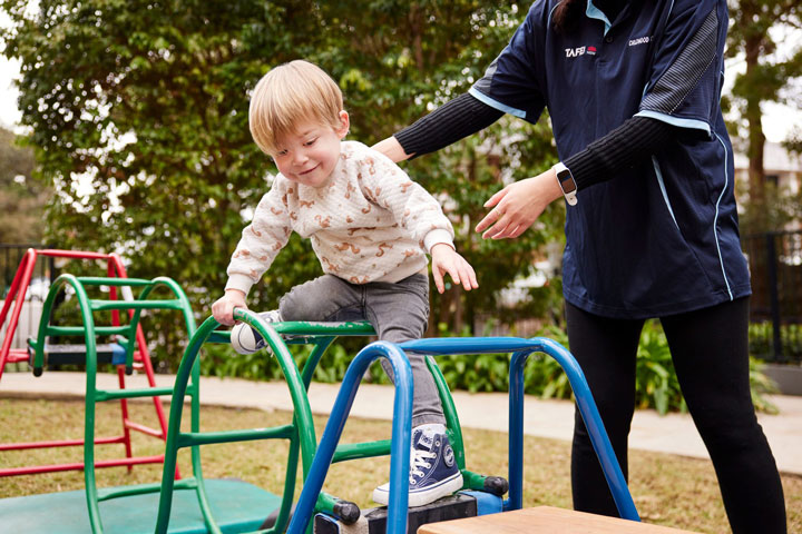 A toddler plays in a colourful outdoor play area, exploring a metal climbing frame with a big smile on their face. A TAFE NSW staff member supervises nearby, ensuring a safe environment.