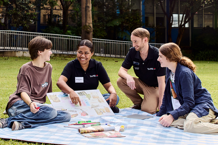 Group of students and advisors having a discussion while sitting on a picnic blanket outdoors on a campus lawn.