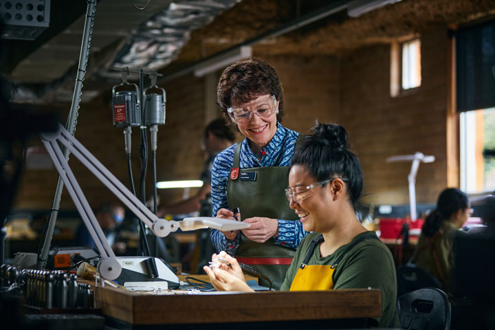 A teacher and student collaborate in a workshop environment. The teacher is smiling and offering guidance while the student uses her hands and works with tools under a light. The setting suggests this is a practical learning task such as jewellery making or metalwork.