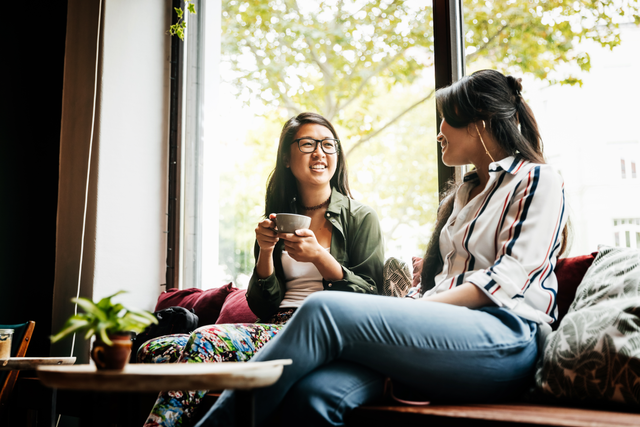 Two women are talking in front of a bright sun-filled window. It appears to be a relaxed atmosphere with one drinking a hot beverage while engaging in conversation with the other. 