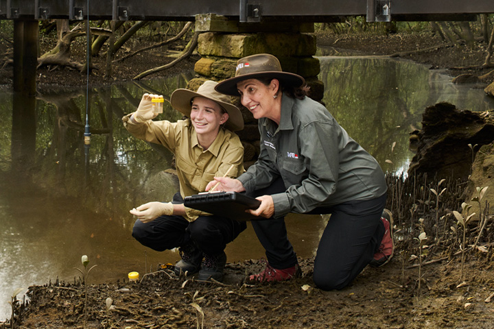 A teacher and student bend down at the edge of a creek, carefully collecting water samples in containers for a science experiment.