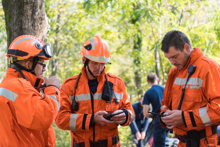 Three men in bright, orange, hi-vis uniforms and hard-hats are standing and programming their walkie-talkies. They are surrounded by bush and established trees, as other workers in the background put on their uniforms.