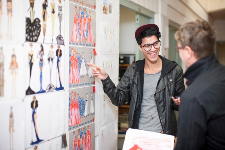 A young man is pointing to some fashion design sketches that are pinned up on the wall. He is speaking to someone and is smiling. He is wearing a leather jacket, grey t-shirt, a cap and some bold-framed glasses.