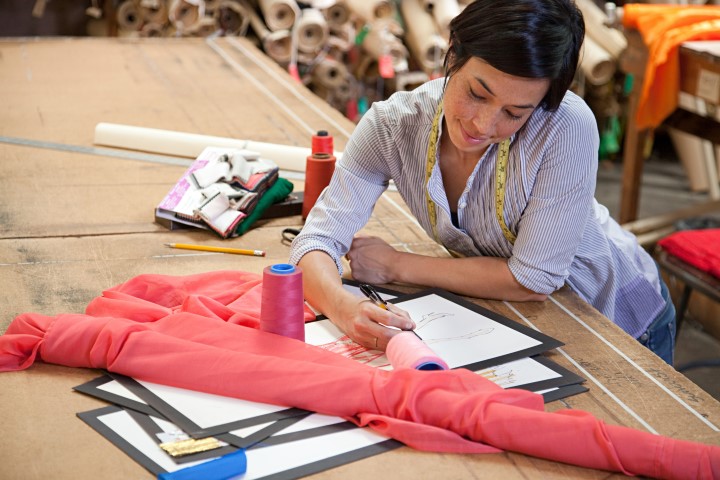 A woman is leaning on a wooden table, working on some fashion design sketches. A ream of material and a spool of wool by her side, as well as a pencil and other design items. She has a measuring tape around her neck and looks pleased.