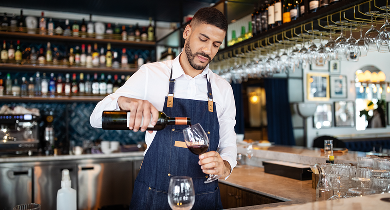 A young, well-groomed young man in a modern, tidy and well-stocked bar carefully pours a glass of red wine from a bottle. Another empty glass rests on the bar. He wears a white-collared shirt and a clean, navy-blue apron. His gaze is concentrated. 