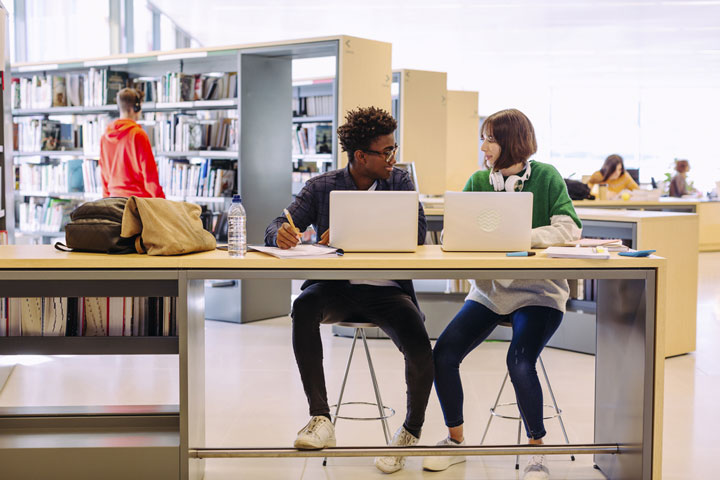 Two students sit in the library with laptops in front of them, reviewing TAFE NSW policies and smiling at each other.