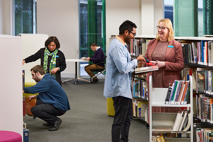 TAFE NSW library with a man seated on the background and library staff helping people who are browsing shelves and accessing resources.