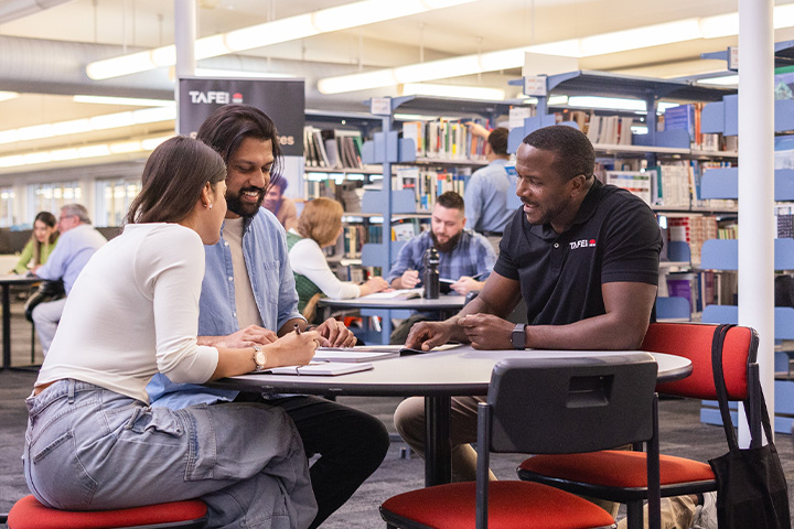 A group of students gathered around their teacher in a brightly lit public library. The students are engaged in a collaborative learning activity, some looking at books and others working at tables.