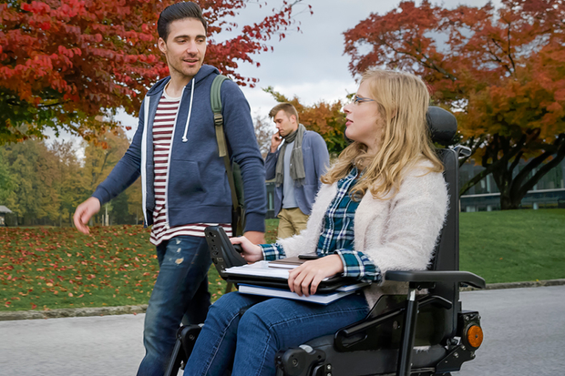 A female student in a motorised wheelchair is talking to a male student walking beside her on a campus path.