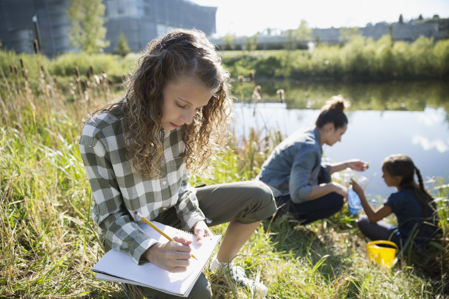An adult and two students collect water samples on a sunny, grassy riverside, in an urban area.