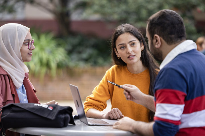 A group of three students are sitting at a table, engaged in conversation as they work on a laptop together.