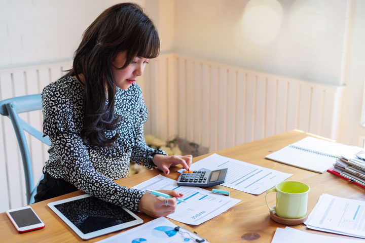 A woman sits at a desk with her work in front of her. She holds a pen in one hand and a calculator in the other. She has papers, an iPad, iPhone and a coffee mug on her desk. 