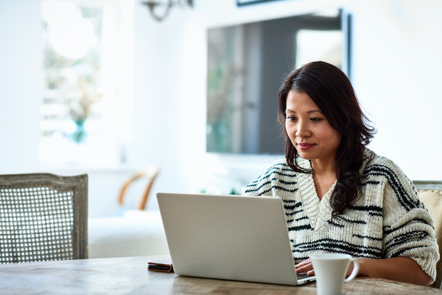 A woman working on a laptop at a wooden table in a bright living room, with a coffee mug nearby.