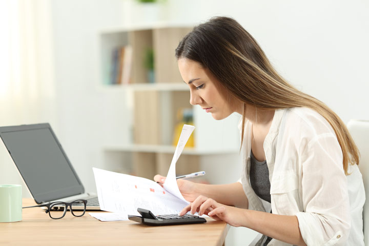 A women sits at her desk looking at a piece of paper and her calculator. 