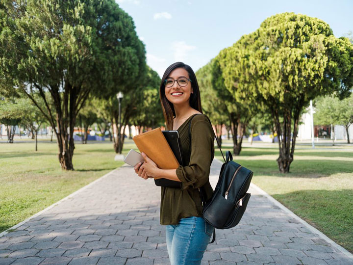 A young woman with glasses, wearing a green blouse and jeans, stands on a tree-lined path holding a folder, laptop, and smartphone, smiling confidently with a backpack over her shoulder.