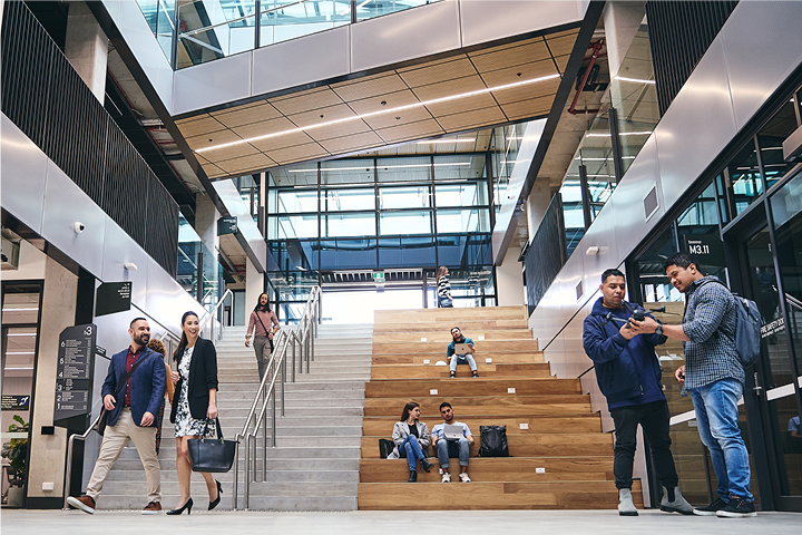 Modern, light filled campus atrium with people in transit, standing in conversation and some seated on the bench side of a wide stairway.