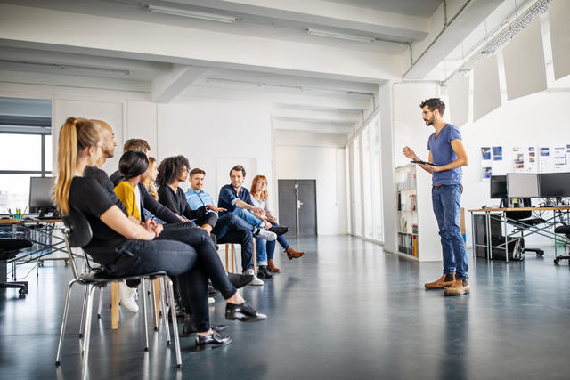The image shows a group of people sitting in chairs in a semi-circle in a spacious, modern, open office environment. They are attentively listening to a man standing in front of them, who appears to be leading a discussion or presentation. The man is holding a tablet and gesturing with his hand. The office is bright, with large windows, sleek furniture, and a clean design.