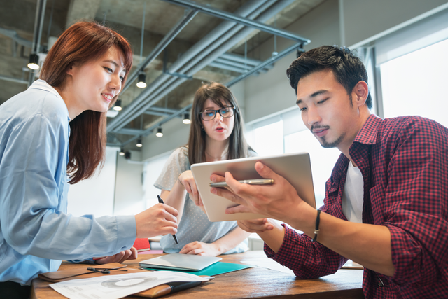 Three colleagues collaborating in a modern office, discussing and reviewing information on a tablet at a wooden table.