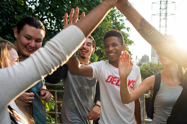 A multicultural group of students smiling standing in a circle. They are reaching up, meeting their hands in the centre in celebration.