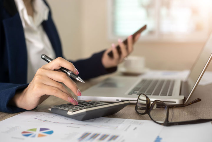 A close-up of a person’s hands calculating payment. One hand types on a calculator while holding a pen, and the other holds a mobile phone.