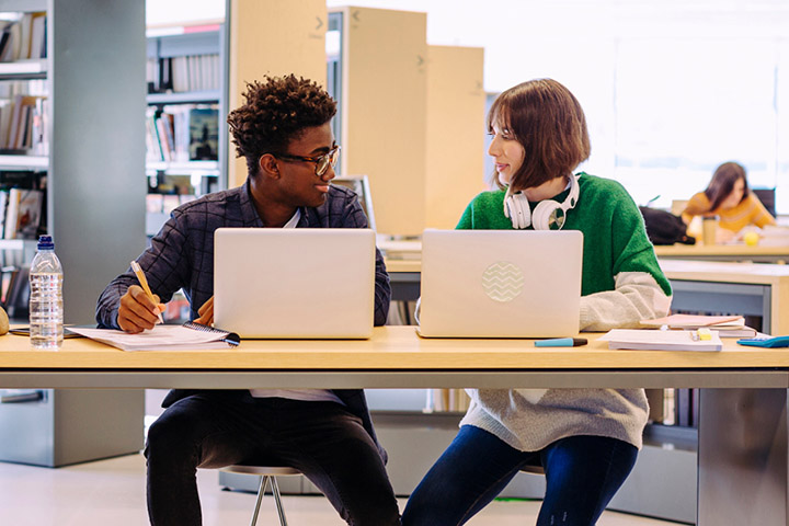 Two students sit in the library with laptops in front of them, talking with each other.