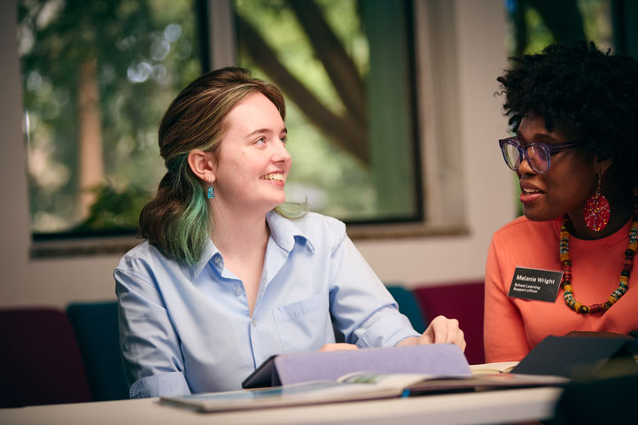A student with blue streaks in her hair sits next to a student learning support officer. They are using a tablet between them.
