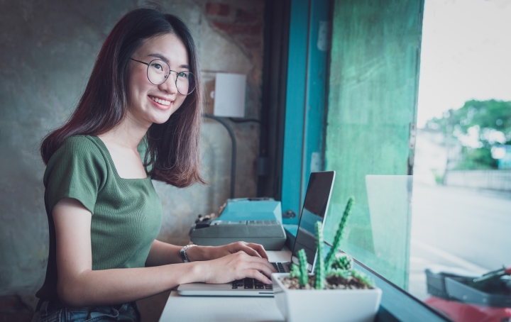 A woman looks happy while sitting at her desk using her laptop. Her desk faces out to a street-facing window, and a cactus is on the desk in the foreground. 