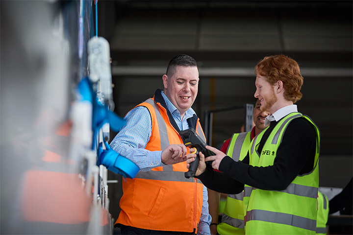 Three people in high-visibility vests on a factory floor. A logistics teacher, wearing an orange vest, points out features of equipment to two students wearing yellow vests.