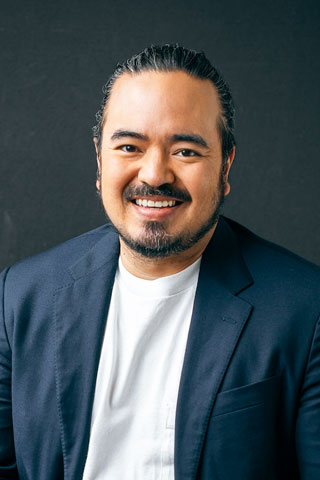 The image shows Adam Liaw in a blazer worn over a white T-shirt. He has his hair pulled back and some facial hair. He is smiling warmly at the camera.