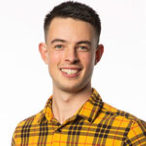 A headshot image of Aidan Williams smiling at the camera. He is wearing a bright yellow and black tartan shirt. 