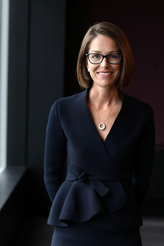 The image shows Alison Mirams in a professional and stylish black outfit. She stands with her hands behind her back and is smiling. She has shoulder length brown hair and is wearing glasses and a necklace. 