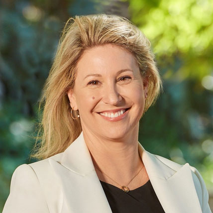 The image shows Eva Hanley smiling towards the camera wearing a white blazer won over a black top. She is wearing fine gold jewellery and has long blonde hair behind her shoulders.