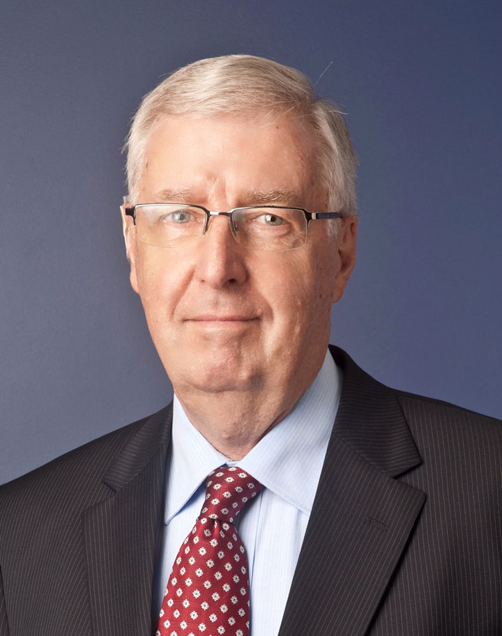 The image shows Geoffrey Newcombe a business suit, wearing a white shirt and a patterned red tie. He has short, grey hair and is wearing glasses.