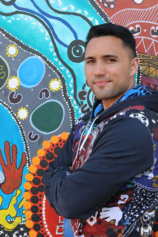 The image shows Isaiah Dawe standing sideways, facing the camera with his arms crossed. He is stands against a traditional Aboriginal painted background, wearing a jumper that matches. 