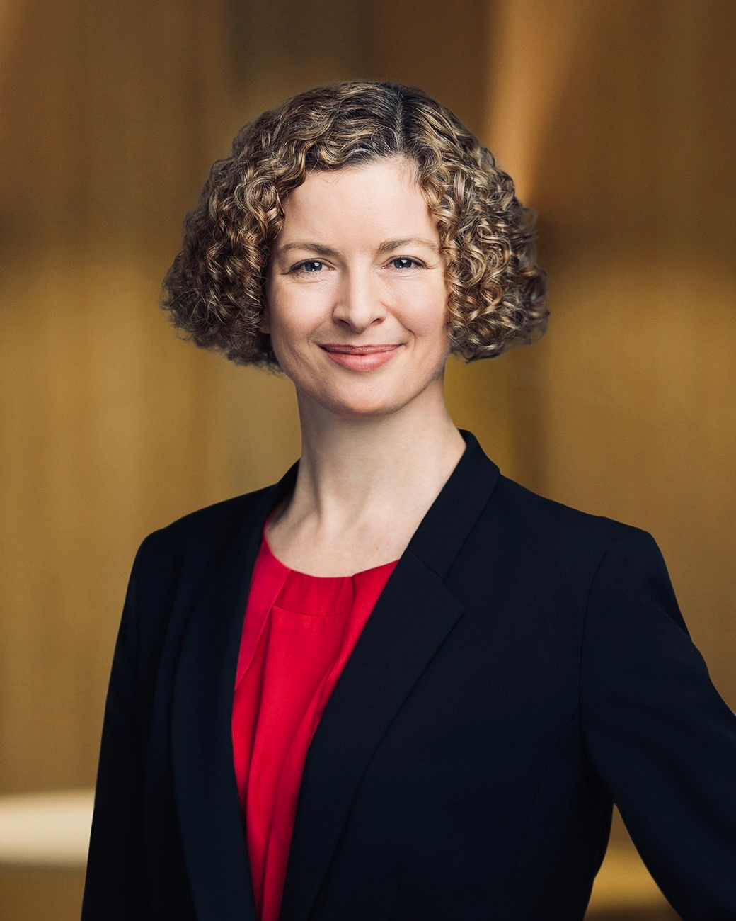 A headshot of Jessica Tilley, smiling and looking directly at the camera. She has short, curly bronde hair, and is wearing a black blazer over a red top.