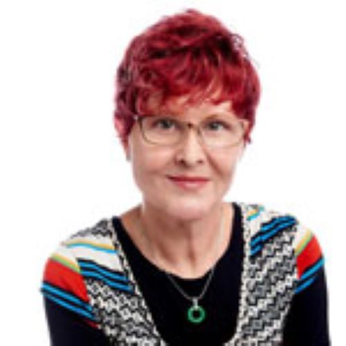 A headshot image of Lydia Kullik smiling at the camera. She is wearing a bright coloured patterned shirt over a black top, and has bright red hair and glasses.