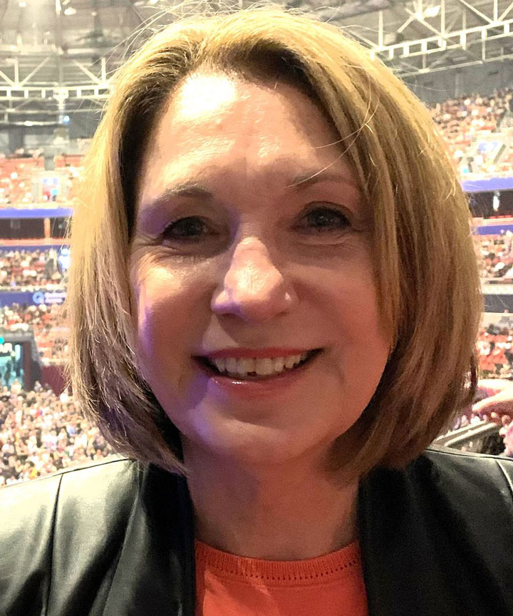 The image shows Mary Ann O’Loughlin up close. She's wearing a black jacket with a red shirt underneath. She has a short, light brown bob haircut and is smiling.