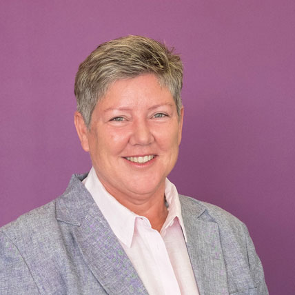 The image shows Megan Callinan in a grey blazer worn over a white collared shirt . She has short, light hair and is smiling.