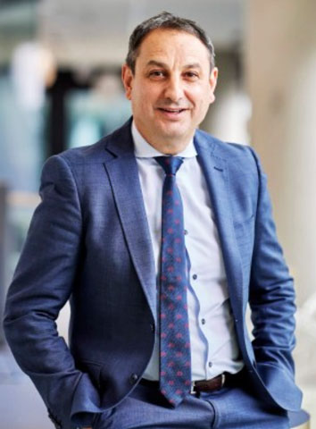 The image shows Murat Dizdar in a dark blue business suit, wearing a patterned blue tie. He has short hair and is smiling with his hands in his pockets.
